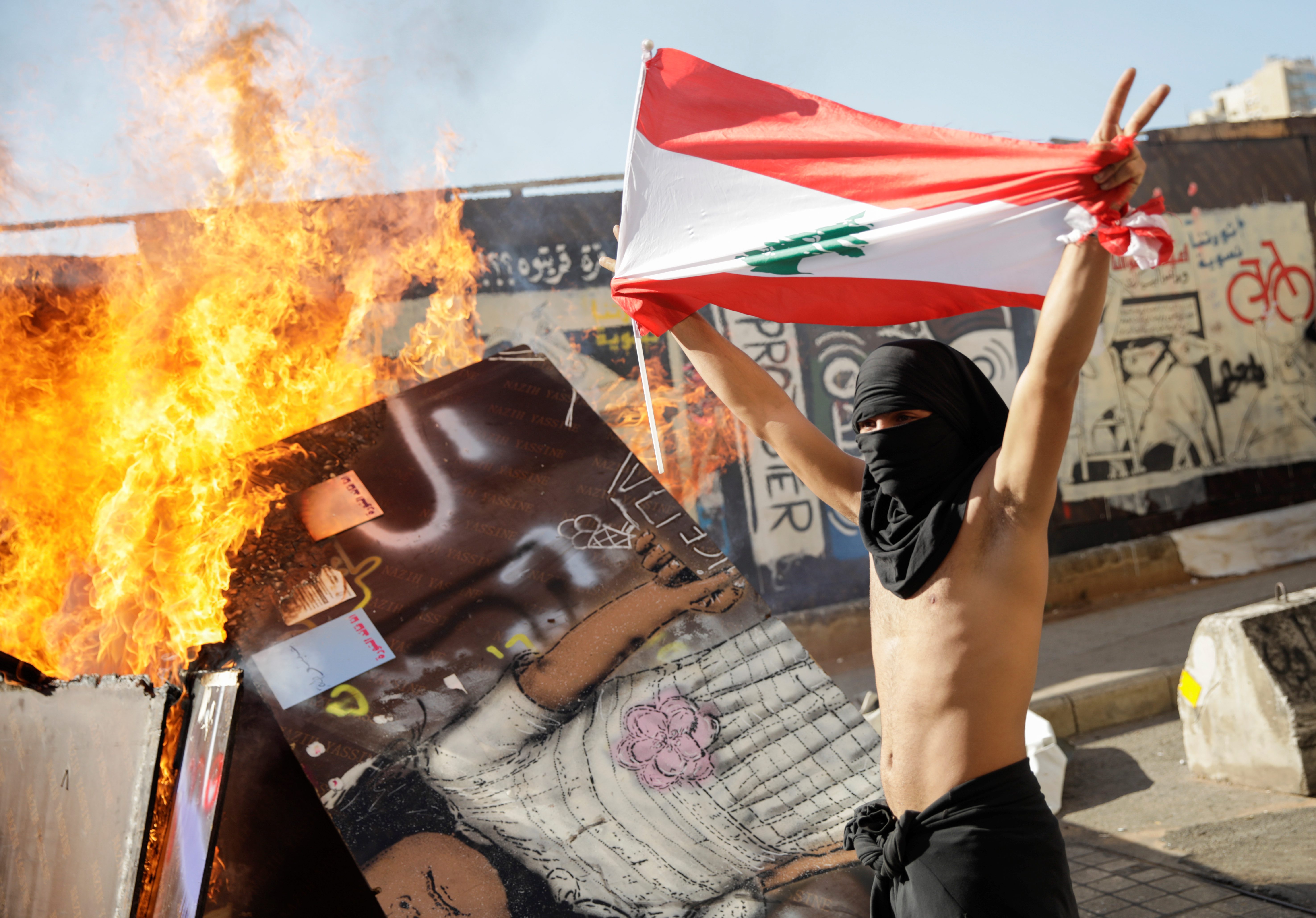 متظاهر ملثم يحمل علمًا لبنانيًا يتخطى حاجزًا محترقًا خلال الاحتجاجات المستمرة/ رويترز
